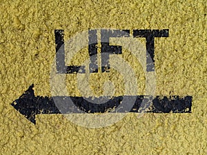 Word LIFT and arrow painted in black on yellow wall pointing at the direction of the lift / elevator