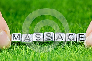 Word MASSAGE written in metal letters laid on grass and held between the fingers of a woman. Concept of wellness background