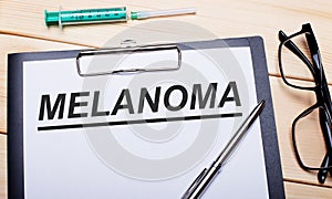 The word MELANOMA is written on a white piece of paper next to black-rimmed glasses, a pen and a syringe. Medical concept