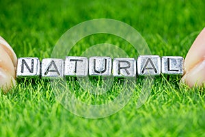 Word NATURAL written in metal letters laid on grass and held between the fingers of a woman. Concept of wellness background