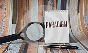 The word paradigm is written on a white notebook, near a pen and a magnifying glass