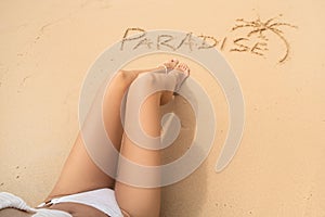 Word paradise and one palm on the sandy beach near the women`s legs. Summer vacation concept. Realax