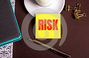 The word RISK written on yellow paper on a brown background near a coffee cup and diaries