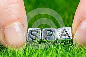 Word SPA written in metal letters laid on grass and held between the fingers of a woman. Concept of wellness background