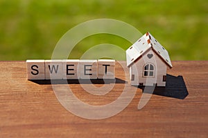Word sweet of wooden letters and small toy wooden house on brown surface, green grass in background. Concept sweet home, mortgage