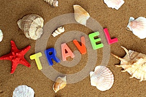 The word travel surrounded by seashells is written in colored letters on the sand.