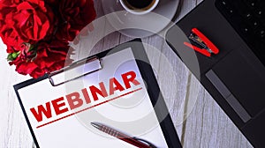 The word WEBINAR is written in red on a white notepad near a laptop, coffee, red roses and a pen