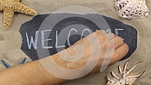 Word Welcome, written on a chalkboard among seashells and starfishes lying on the sand. Handwritten words.