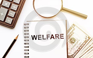 The word WELFARE written on notebook on table with calculatir, magnifier, pencil and dollars