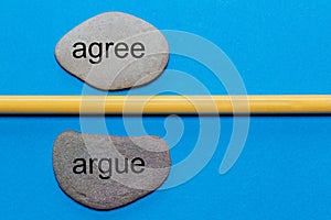 The words agree and argue are written on natural polished stones separated by a yellow pencil. The background is isolated in blue