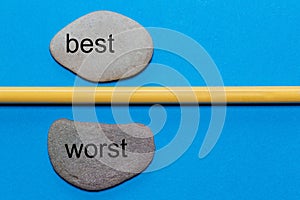 The words best and worst are written on natural smooth stones separated by a yellow pencil. The background is isolated in blue and