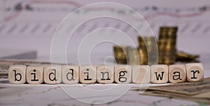 Words BIDDING WAR composed of wooden letter. Stacks of coins in the background