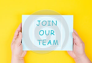 The words join our team are standing on a paper, yellow colored background, hands hold message