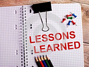 The words LESSONS LEARNED is written in a notebook near multi-colored pencils and buttons on a wooden background