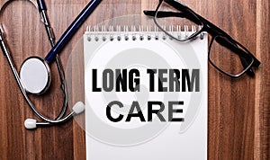 The words LONG TERM CARE is written on white paper on a wooden background near a stethoscope and black-framed glasses. Medical