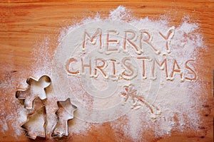 Words `Merry Christmas` written in spread flour and a variety of cookie cutters on a wooden chopboard