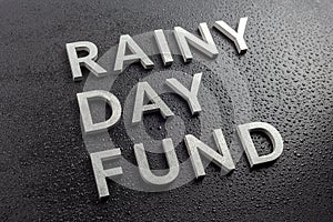 The words rainy day fund laid with silver metal letters on black background, covered with water drops, diagonal perspective,