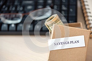 Words savings plan print on pieces of paper, money banknotes dollars in craft box, glasses and on table