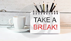 The words TAKE A BREAK is written in a white notepad near a white cup of coffee on a light background