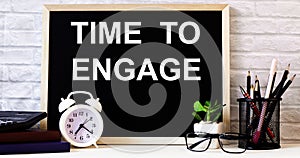 The words TIME TO ENGAGE is written on the chalkboard next to the white alarm clock, glasses, potted plant, and pencils in a stand
