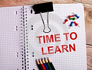 The words TIME TO LEARN is written in a notebook near multi-colored pencils and buttons on a wooden background