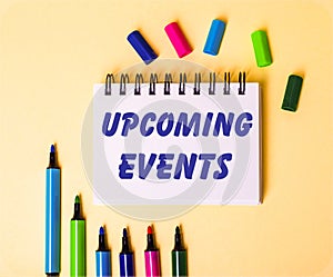 The words UPCOOMING EVENTS written in a white notebook on a beige background near multi-colored markers