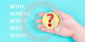 The words who, what, why, when, where are standing on a blue colored background, hand is holding a question mark