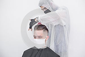 The work of the barber during the coronavirus, the hairdresser trim the client in a mask and a protective suit, quarantine