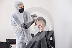 The work of the barber during the coronavirus, the hairdresser trim the client in a mask and a protective suit, quarantine