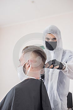 The work of the barber during the coronavirus, the hairdresser trim the client in a mask and a protective suit, quarantine