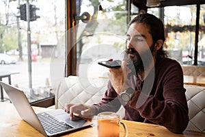 Work from cafe. Young hipster man talking on mobile phone, using voice assistant. Bearded guy with long hair sitting at