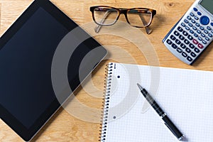 Work desk with tablet, glasses, calculator and notepad on wooden