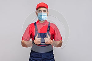 Work are done! Portrait of young man with surgical medical mask in blue overall, red t-shirt , cap, standing and showing thumps up