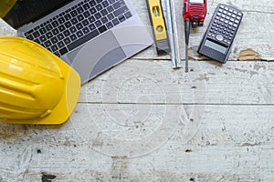 work environment with a laptop, a yellow hard hat, calculator, level, screwdriver, and folding ruler on a well-used wooden surface