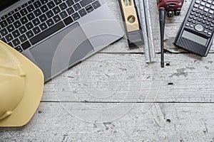 work environment with a laptop, a yellow hard hat, calculator, level, screwdriver, and folding ruler on a well-used wooden surface