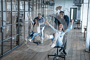 Work hard play hard. Four young cheerful business people in smart casual wear having fun while racing on office chairs and smiling