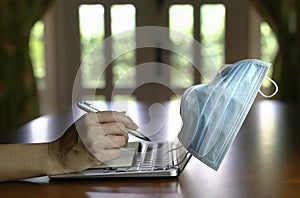 Work at home during coronavirus pandemic. Focus on face mask with woman`s hands on computer laptop and notepad on dining table