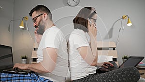 Work at home, freelance concept. Husband and wife sitting on bed in bedroom, back to back, while working, typing on