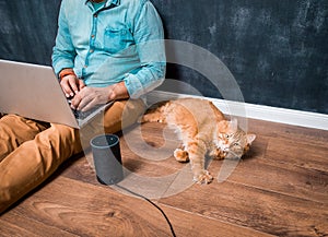 Work from home with funny lazy red cat pet. Man sitting on laminate wooden floor with laptop computer and smart speaker