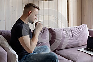 Work at home online. Young man with short stylish haircut in black T-shirt and jeans is sitting on cozy sofa and