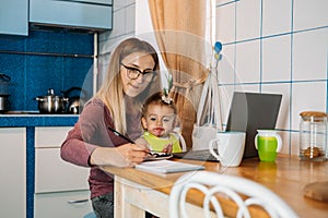 Work from home. Young mother with baby girl working at home using laptop on kitchen background Young woman feeding her baby,