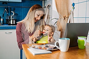 Work from home. Young mother with baby girl working at home using laptop on kitchen background Young woman feeding her baby,