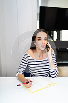 Work at home. Young woman uses a laptop to work in the kitchen writes something in a notebook with a pen and speaks on the phone