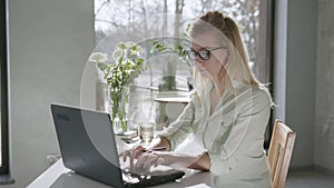 Work online, beautiful attractive blonde woman works at laptop in glasses for vision and takes notes in notebook, then