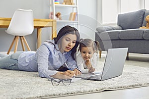Work online. Woman working from home using laptop computer lying on livingroom carpet with curious daughter.