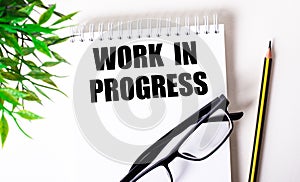 WORK IN PROGRESS is written in a white notebook next to a pencil, black-framed glasses and a green plant