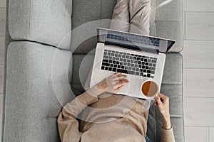 Work remotely from home: female reading reports and emails on laptop lying with cup of tea on couch