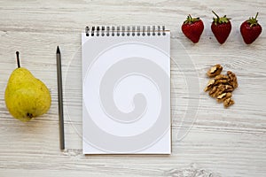 Work space for healthy person: blank notepad, pencil, walnuts, strawberries and pear on a white wooden background.