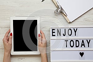 Work space with tablet, notepad and `Enjoy today` word on lightbox over white wooden background, top view. Female hands hold tab