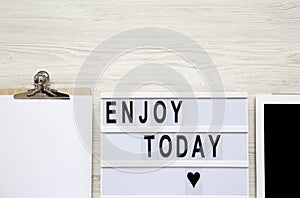 Work space with tablet, notepad and `Enjoy today` word on lightbox over white wooden surface, top view. From above, flat-lay, ov